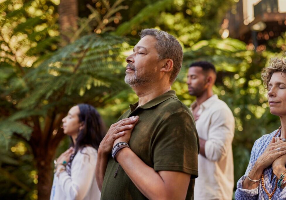 Group of multiethnic people standing outdoors with eyes closed and hands on their hearts during a meditation session. Multiracial adults practicing gratitude and mindfulness together. Mature latin man connecting to inner peace and presence at wellness retreat center in nature.